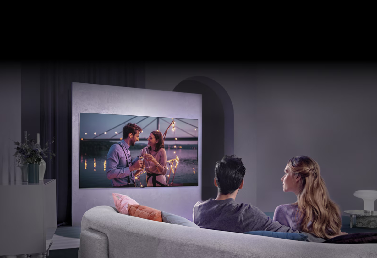 Απεικονίζεται η τηλεόραση κρεμασμένη στον τοίχο σαλονιού και δύο άνθρωποι να παρακολουθούν